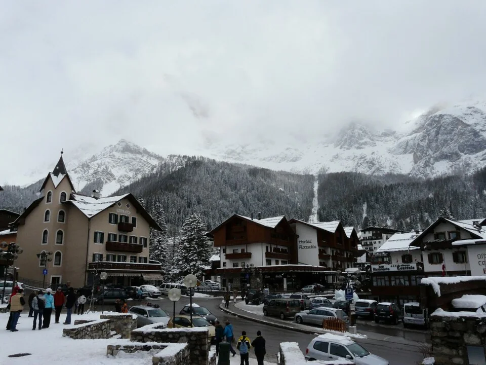 Snow-covered buildings in the centre of San Martino di Castrozza beneath forested slopes and rocky peaks.