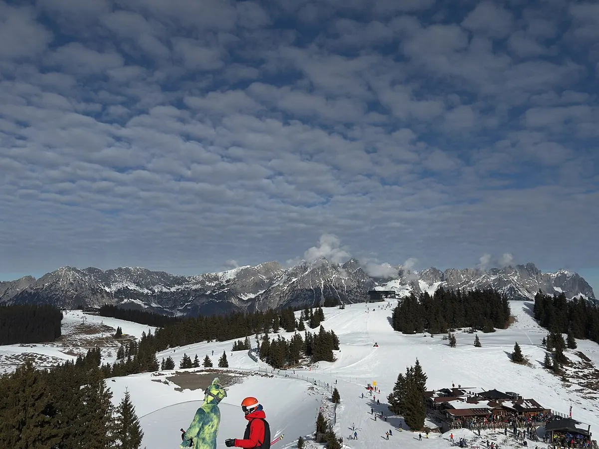Panoramic view of SkiWelt Wilder Kaiser-Brixental ski resort in the Austrian Alps. Two skiers/snowboarders visible in the foreground with dramatic altocumulus cloud formations over snow-covered mountains.